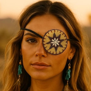 Puede incluir: Un parche en el ojo con cuentas con un diseño de estrella en tonos dorados, marrones y blancos. Se sujeta con una banda elástica negra. La persona también lleva pendientes turquesas.