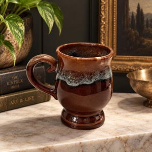 May include: A ceramic mug with a glossy finish featuring a deep brown base and a lighter brown upper section separated by a textured, mottled white and blue drip glaze pattern. The mug has a curved handle and a pedestal base with a rounded body.