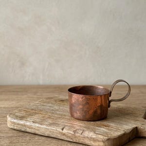 May include: A small, hammered copper cup with a rounded handle sits on a rustic wooden cutting board. The cup has a rich, reddish-brown color and a textured surface. The cutting board is light brown with visible wood grain.