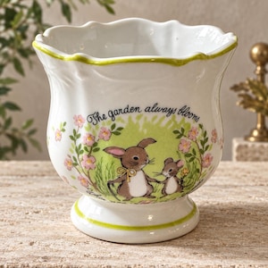 May include: A white ceramic planter with a scalloped rim and a green painted edge. The front features a graphic of a brown rabbit and a small mouse in a green field with pink flowers and the text, The garden always blooms, in black script.