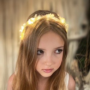 May include: A young girl wearing a white flower crown with small white lights. The crown is made of fabric flowers and has a delicate, feminine look.