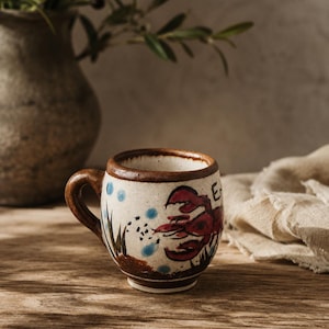 May include: A small, off-white ceramic mug with a brown rim and handle. The exterior features a hand-painted red lobster, green seaweed, and blue bubbles. The surface has a speckled texture, and the base is slightly tapered with a brown painted band.