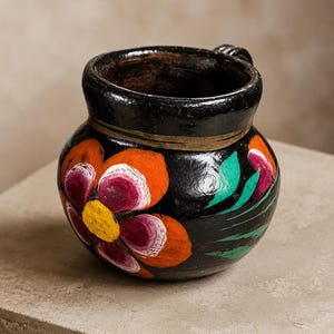 May include: A black ceramic mug with a rounded body and a wide rim. The exterior features hand-painted floral motifs in vibrant orange, magenta, yellow, and green. A single handle is visible on the right side, and a thin horizontal line circles the neck.