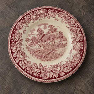 May include: A round ceramic plate featuring a cream-colored base with a deep red transferware pattern. The rim is decorated with a dense border of roses and leaves, while the center depicts a rural landscape with a cottage and trees.