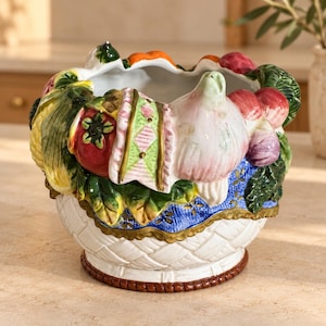 May include: A ceramic bowl featuring a white woven-texture base and a rim decorated with sculpted, hand-painted vegetables and fruits, including a red tomato, a pale garlic bulb, and green leaves, accented by a blue and gold patterned border.