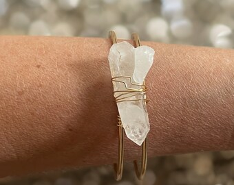 May include: A gold wire wrapped bracelet with a clear quartz crystal. The bracelet is on a person's wrist.