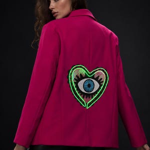 May include: A vibrant magenta blazer featuring a large, embroidered heart-shaped patch on the back. The patch displays a central blue eye motif surrounded by sequins and a bright neon green outline, creating a bold, contrasting visual element.