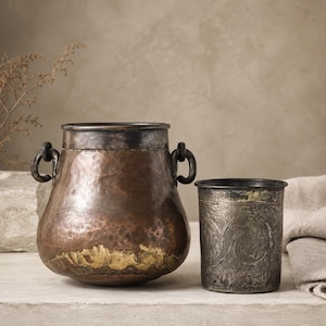 May include: A hammered copper pot with two dark metal side handles and a dark rim, positioned next to a cylindrical metal cup featuring an intricate embossed floral pattern and a dark, weathered finish on the upper and lower edges.