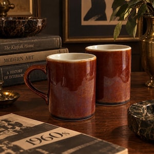 May include: Two cylindrical ceramic mugs with a glossy, deep brown glaze that transitions to a darker tone at the base. Each mug features a smooth, white interior and a sturdy handle on one side, showcasing a reflective finish on the exterior surface.