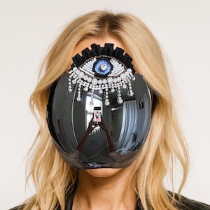 May include: A large, reflective visor with a decorative eye design. The eye is embellished with white beads, pearls, and a blue center, with black trim. The visor reflects the surroundings, including a person holding a phone.