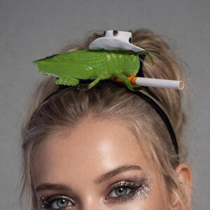 May include: A novelty headband featuring a green grasshopper, a white cowboy hat with black spots, and a cigarette. The grasshopper is perched on a black headband, creating a whimsical and unique accessory.