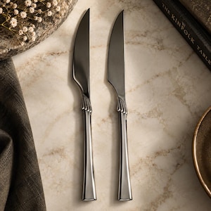 May include: Two silver-toned dinner knives with smooth, polished blades and handles featuring a subtle, fan-shaped decorative detail at the bolster. The knives have a sleek, tapered design with a reflective metallic finish and a solid construction.