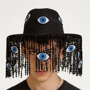 May include: Black fedora hat adorned with multiple embroidered eye patches with blue irises. The hat's brim features a fringe of black sequins and beaded strands, creating a unique and artistic design.