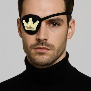 May include: Black eye patch with a gold sequined crown design. The eye patch is worn over one eye. The person is wearing a black turtleneck.