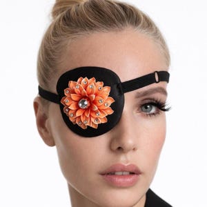 May include: Black eye patch with an orange flower design. The flower has a central jewel and is surrounded by smaller jewels. The eye patch is made of a soft material and has a black elastic strap.