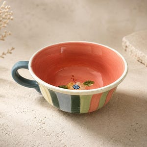May include: A ceramic mug with a wide, shallow bowl shape featuring a coral pink interior and a hand-painted flower at the base. The exterior displays vertical stripes in muted shades of blue, green, and coral, complemented by a solid blue handle.