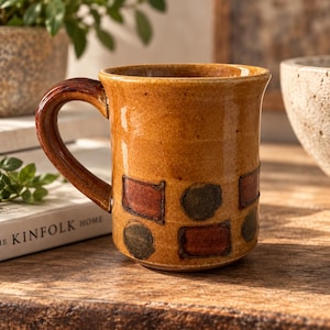 May include: A ceramic mug with a glossy mustard yellow glaze and a dark brown handle. The exterior features a repeating pattern of dark red squares and dark green circles arranged around the lower half of the cylindrical body.