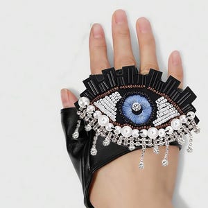 May include: Black leather fingerless glove with an eye design. The eye is embellished with blue, white, and brown beads, pearls, and dangling rhinestones. The glove is a unique fashion accessory.