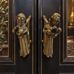 May include: A pair of antique-style brass door handles featuring a sculpted figure holding a child. The metallic gold-toned handles are mounted on a dark black door frame with subtle gold trim, showcasing intricate details on the figures' clothing.