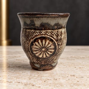 May include: A ceramic cup featuring a dark brown and beige mottled glaze with a prominent circular medallion containing a stylized flower pattern. The body of the cup is decorated with a repeating zigzag texture, and the rim has a dark brown finish.