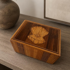 May include: A rectangular wooden box featuring a dark brown wood grain finish with a lighter wood inlay border. The lid displays a centered, laser-engraved illustration of an open seashell with a small sunburst detail in the center of the shell.