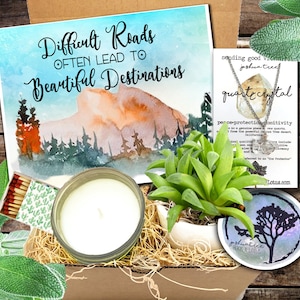 May include: A gift box filled with a variety of items, including a candle, a succulent plant, matches, and a necklace with a clear quartz crystal. The box is decorated with a card that reads "Difficult Roads Often Lead To Beautiful Destinations".
