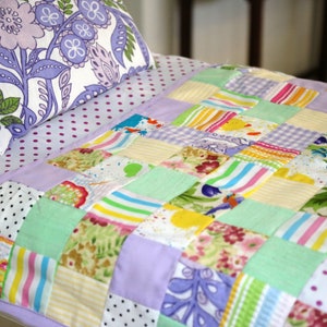 May include: A patchwork quilt featuring a mix of colourful fabrics, including stripes, polka dots and floral patterns. The quilt is square shaped and is laid out on a bed with a purple and white polka dot sheet.