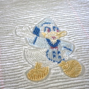 May include: A white, fluffy, striped baby blanket with a blue and yellow embroidered cartoon duck wearing a sailor hat.