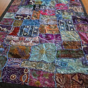 May include: A colourful patchwork textile with a black border. The textile is made up of many fabric squares in shades of blue, purple, pink, and gold. Some squares have embroidered designs and embellishments, creating a textured and decorative piece.