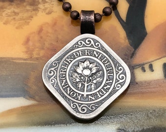 May include: A silver pendant necklace with a square pendant featuring a stylized tree with leaves and a fruit. The pendant is inscribed with the text "KONINKRIJK DER NEDERLANDEN".