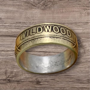 May include: A gold-colored ring with the word "WILDWOOD" embossed around the band. The ring has a brushed metal interior and sits on a wooden surface. The ring is a wide band style.