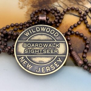 May include: A brass-colored pendant necklace with a round charm that reads "Wildwood Boardwalk Sightseer New Jersey". The charm has a distressed, aged look and is attached to a brown beaded chain.