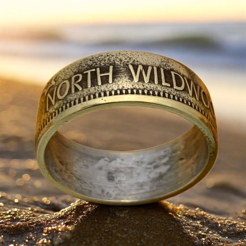 May include: A gold-colored ring with the words "NORTH WILDWOOD" engraved around the top. The ring has a textured, antique appearance and is set against a blurred beach background.