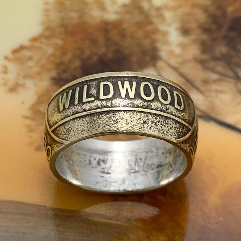 May include: A close-up of a brass-colored ring with the word "WILDWOOD" embossed around the band. The ring has a textured, aged appearance. The background is blurred, with warm tones of brown and gold.