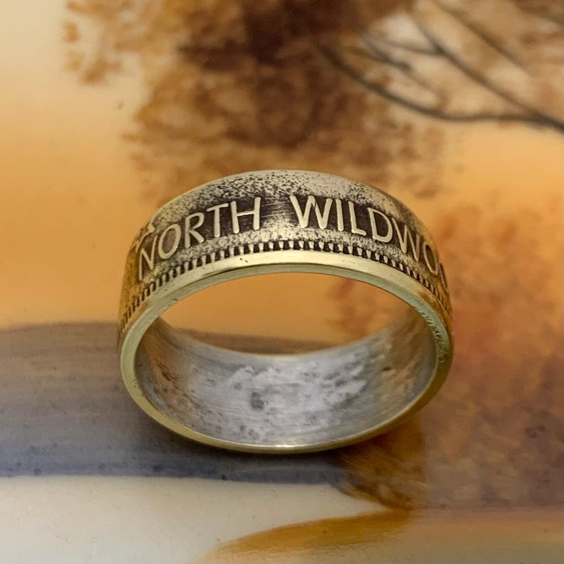 May include: A gold-toned ring with the words "NORTH WILDWOOD" engraved around the band. The ring has a textured, aged appearance, with a contrasting polished interior. The ring is a close-up shot, showcasing the details of the inscription and metalwork.
