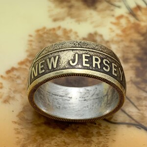 May include: A vintage-style ring with the words "NEW JERSEY" engraved in bold letters. The ring has a two-tone design, with a silver interior and a gold-colored exterior. The ring is a unique piece of jewelry.