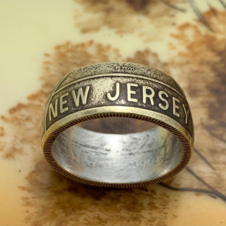 May include: A vintage-style ring with the words "NEW JERSEY" engraved in bold letters. The ring has a two-tone design, with a silver interior and a gold-colored exterior. The ring is a unique piece of jewelry.