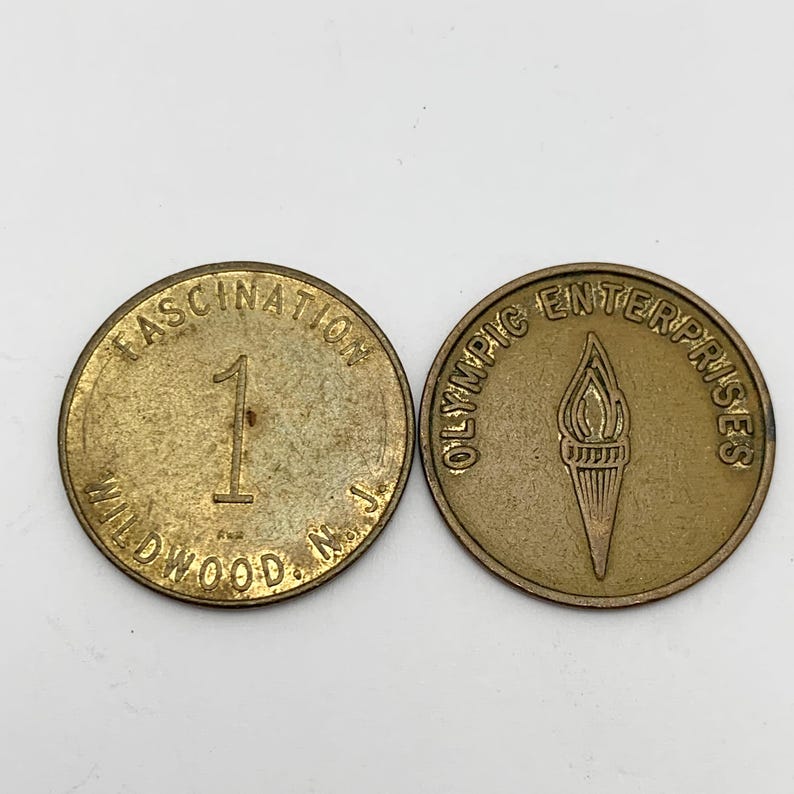 May include: Two gold-colored tokens. One token has the text "OLYMPIC ENTERPRISES" around the edge and a torch design in the center. The other token has the text "FASCINATION WILDWOOD N.J." around the edge and the number "1" in the center.