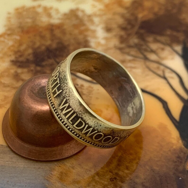 May include: A gold-colored ring with the words "NORTH WILDWOOD" engraved around the band. The ring is resting on a small, copper-colored bowl. The background has a blurred, artistic design.
