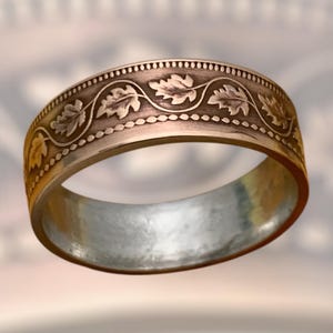 May include: A gold-toned ring with an engraved leaf pattern. The ring features a band of small dots along the top and bottom edges, with a flowing vine and leaf design in between. The interior of the ring has a silver-colored finish.