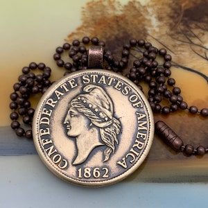 May include: A brass pendant necklace featuring a replica of a Confederate States of America coin from 1862. The coin depicts a bust of Liberty with a Phrygian cap and the inscription "CONFEDERATE STATES OF AMERICA".
