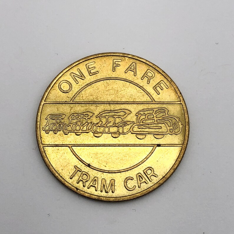 May include: A round, gold-colored token with the words "ONE FARE" and "TRAM CAR" in a circular design. The center features an embossed image of a vintage tram car. The token is likely a vintage transit fare.