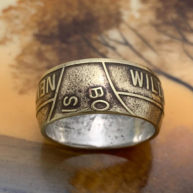 May include: A brass-toned ring with the words "NEW IS BO WILD" engraved around the band. The ring has a vintage, worn appearance, with a silver interior. The ring's design features a geometric pattern.