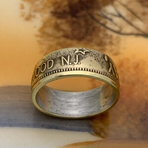 May include: A gold-toned ring with the text "WOOD N.J." and a star design. The ring has a detailed, textured surface and a polished interior. The ring is a piece of jewelry.
