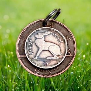 May include: A circular pendant crafted from two layered copper-toned coins. The smaller top coin features an embossed hare and the text "leat reul" with "3d" below. A dark metal split ring is attached to the top for hanging purposes.