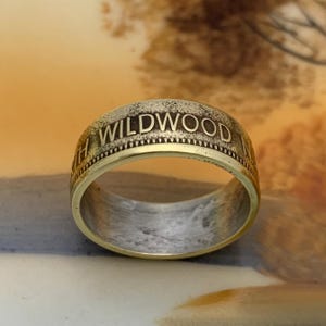 May include: A gold-toned ring with the word "WILDWOOD" engraved around the band. The ring has a two-tone design, with a gold outer band and a silver-colored inner band. The ring is a close-up shot.