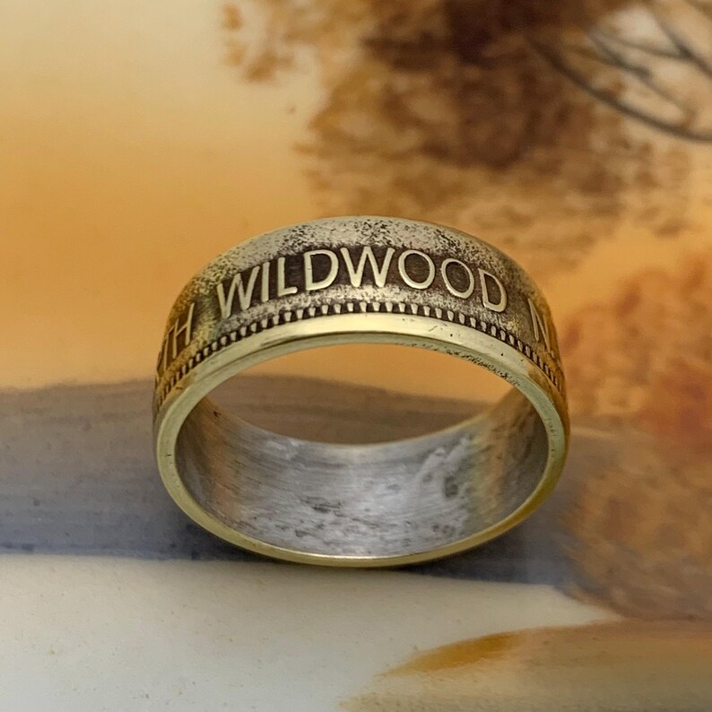 May include: A gold-toned ring with the word "WILDWOOD" engraved around the band. The ring has a two-tone design, with a gold outer band and a silver-colored inner band. The ring is a close-up shot.