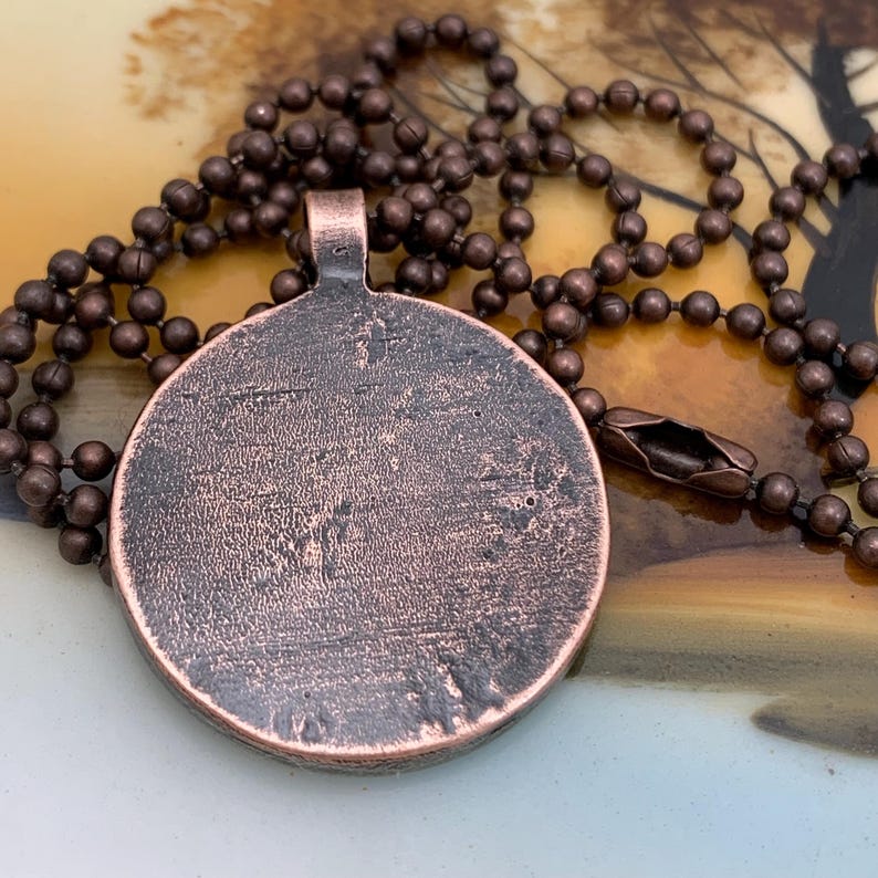 May include: A copper pendant necklace with a round, textured copper charm. The charm has a smooth, shiny surface and is attached to a brown ball chain.