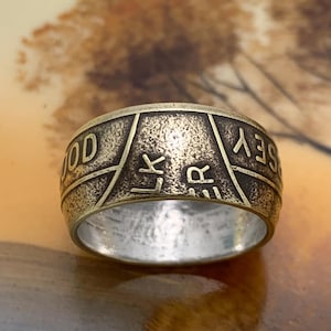 May include: A bronze-colored ring with engraved text. The ring has a textured surface and a wide band. The text includes the words "GOOD LUCK JERSEY". The ring is likely made of metal and has a vintage or antique appearance.