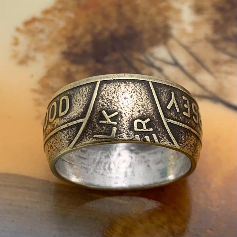 May include: A bronze-colored ring with engraved text. The ring has a textured surface and a wide band. The text includes the words "GOOD LUCK JERSEY". The ring is likely made of metal and has a vintage or antique appearance.